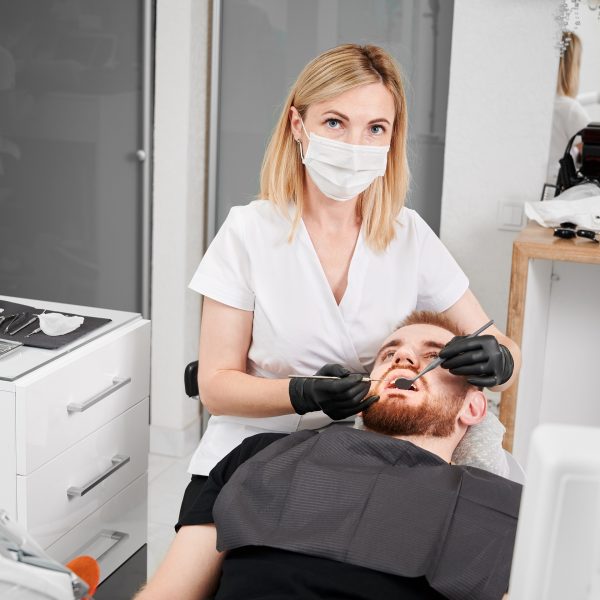 Woman stomatologist in medical mask looking at camera while examining male patient teeth with dental explorer and mirror. Concept of dentistry, stomatology and dental care.